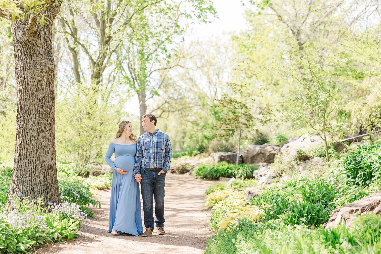A couple walking together on a garden path surrounded by spring blooms and tall trees on a sunny day at Chicago Botanic Garden