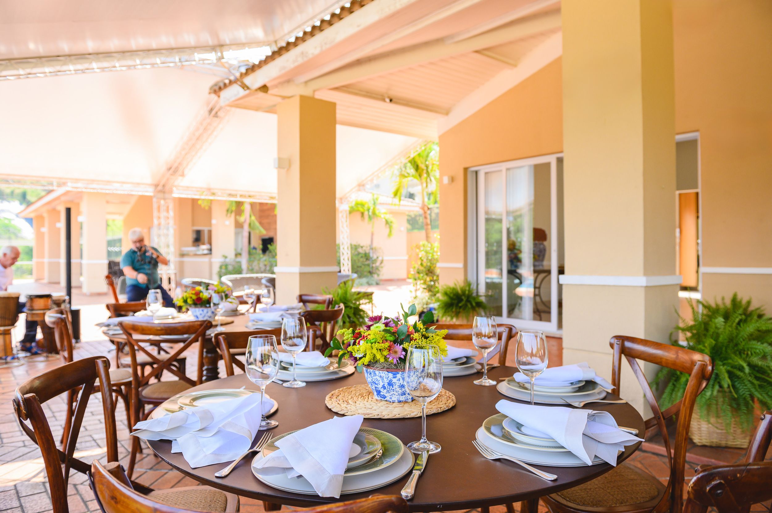 Elegant outdoor dining area with formal table settings, wooden chairs, and cream-colored walls under a covered patio.