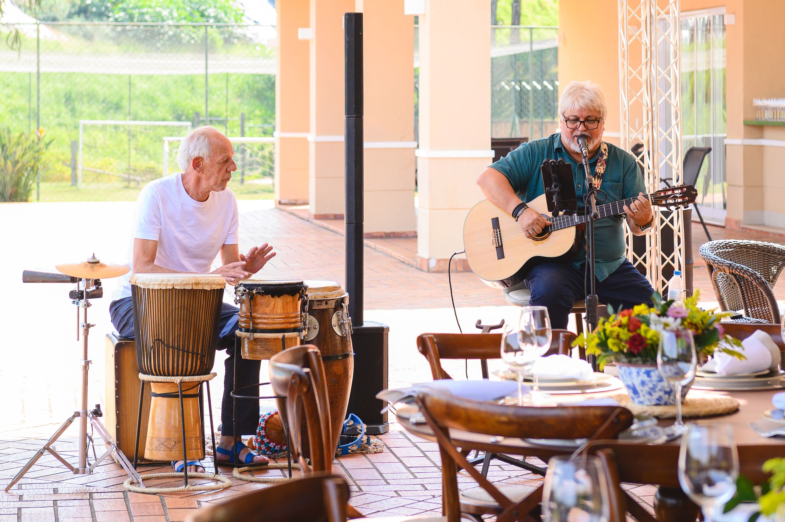 Musicians performing with percussion drums and acoustic guitar on an outdoor patio with dining tables and flowers.