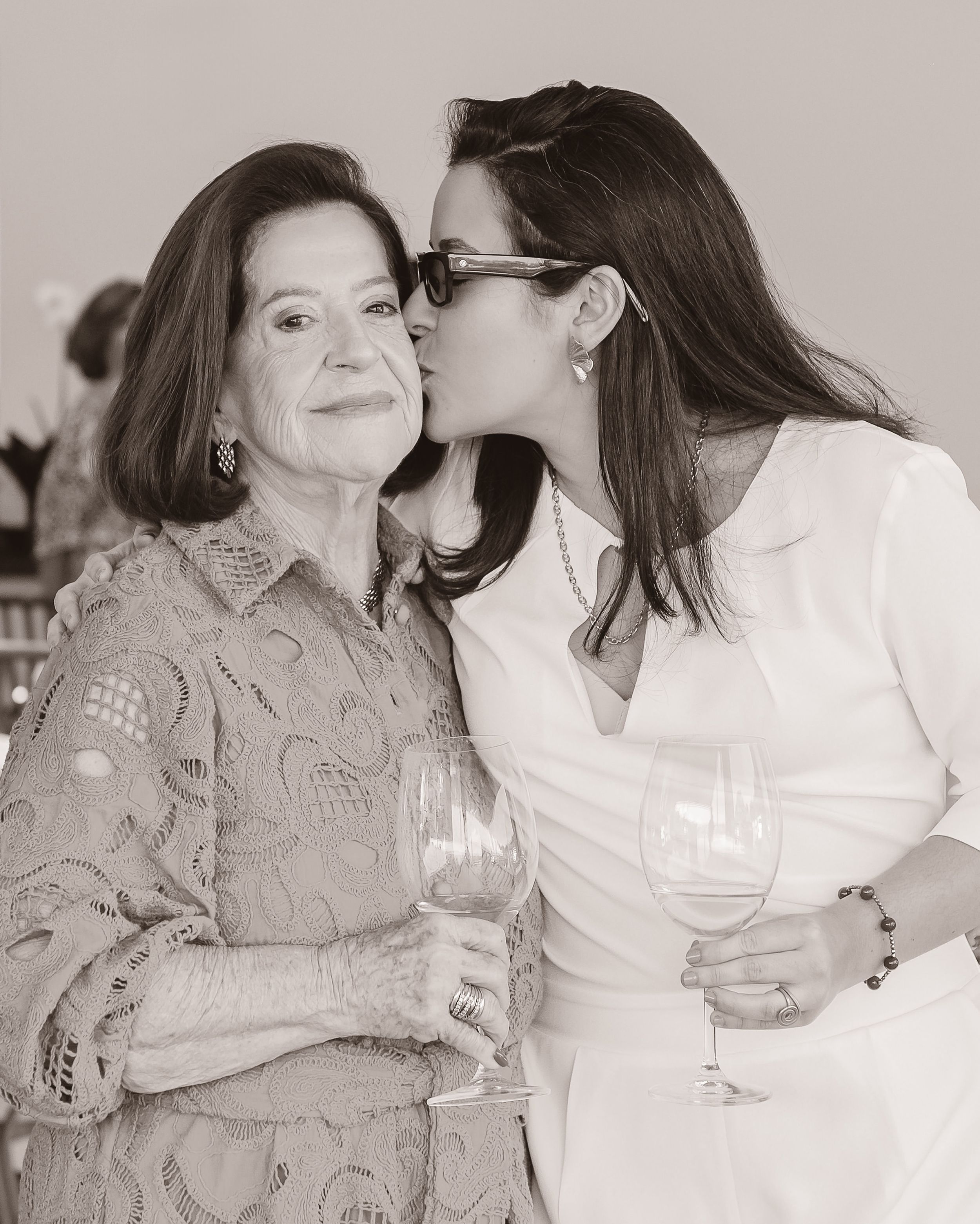 A black and white photo shows two women standing close together holding wine glasses while one gives a kiss on the cheek.