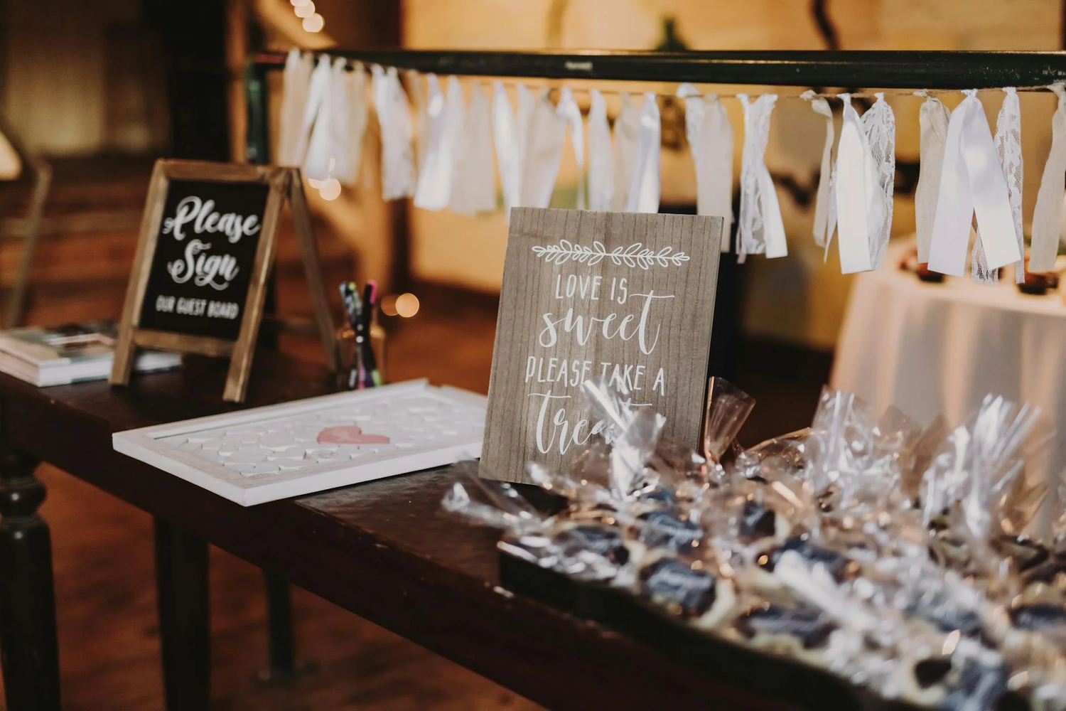 Wedding reception decor with rustic signage and white fabric draping displays event details and favors on dark wooden tables.