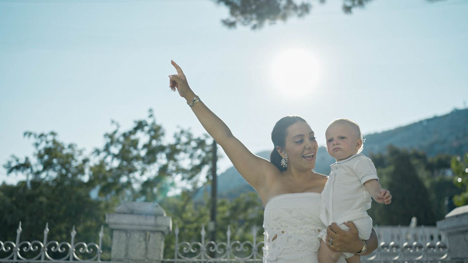 A joyful parent holding a baby and pointing upward outdoors on a bright sunny day with mountains in the background.