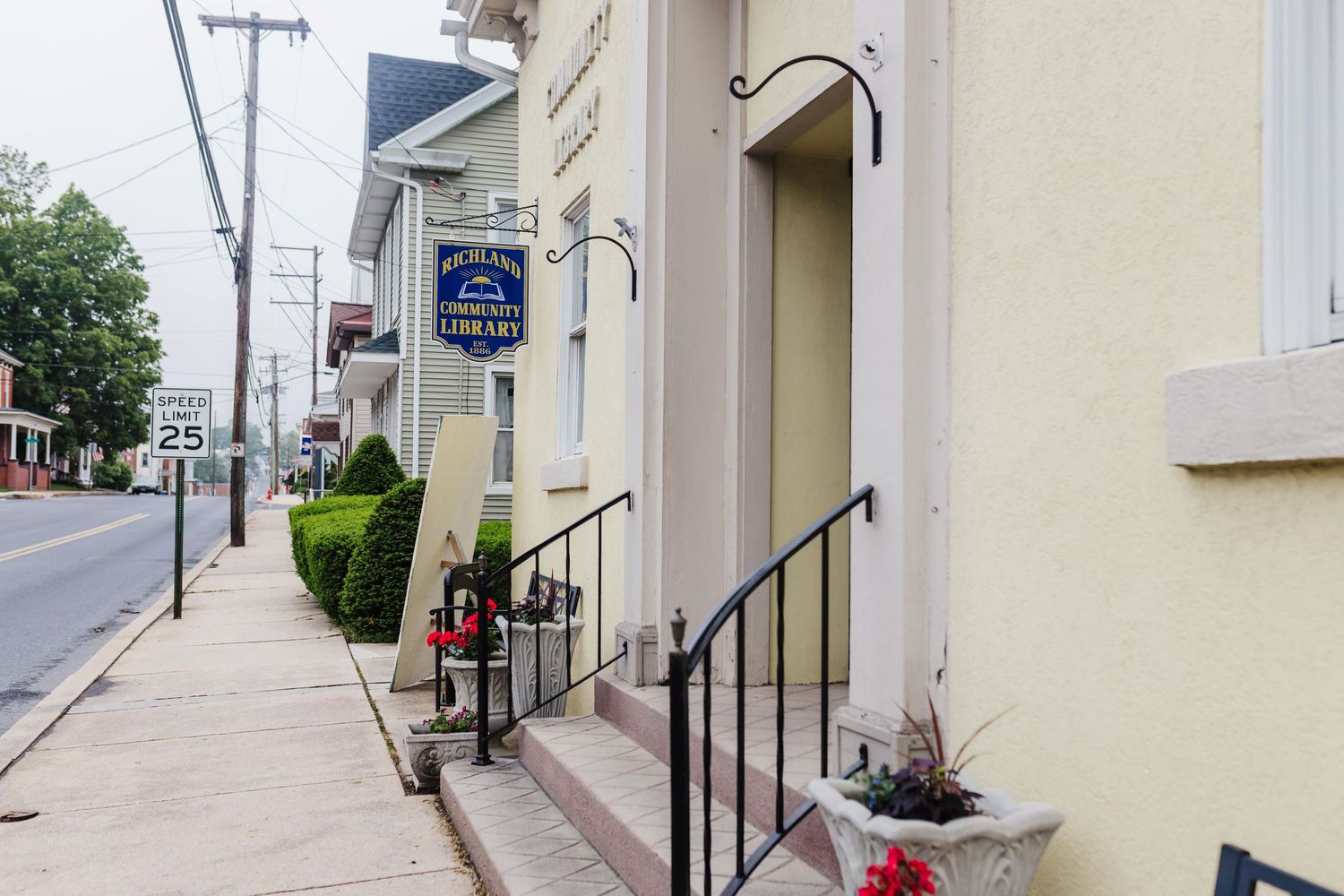 Iron handrails lead up steps to a cream colored building entrance with hanging lamp and iron Richland Community Library sign on Main Street.