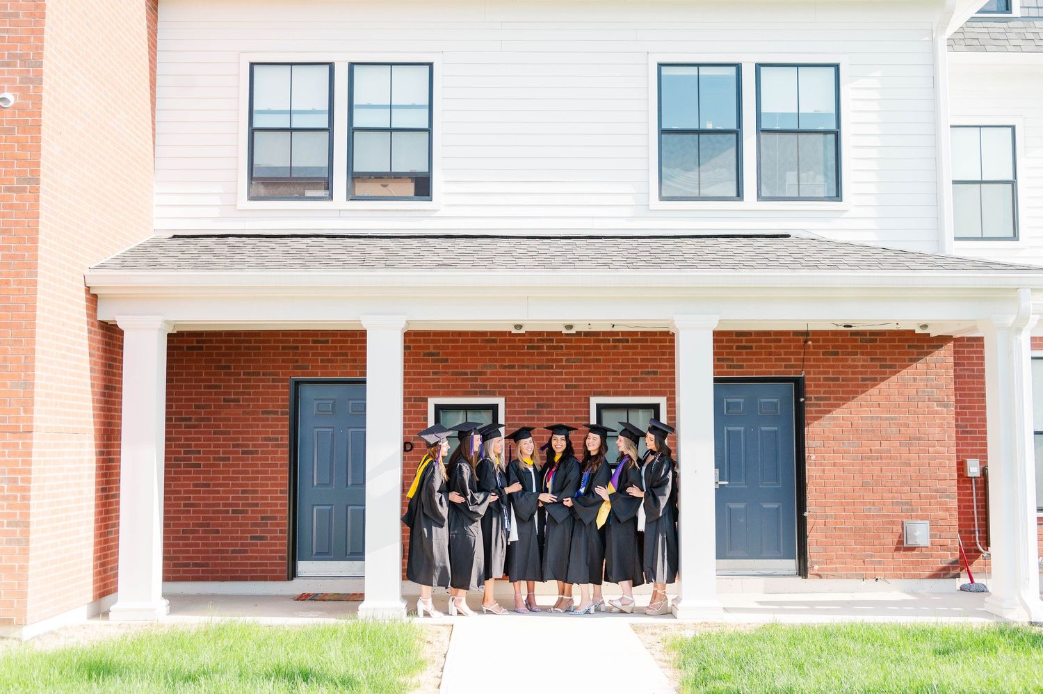 A group of graduates in black caps and gowns pose together on the front porch of a brick building.
