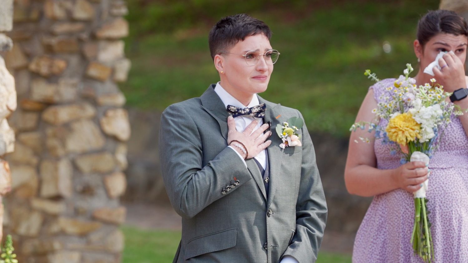 Wedding guest in gray suit wipes tears during emotional outdoor ceremony with stone columns in background.