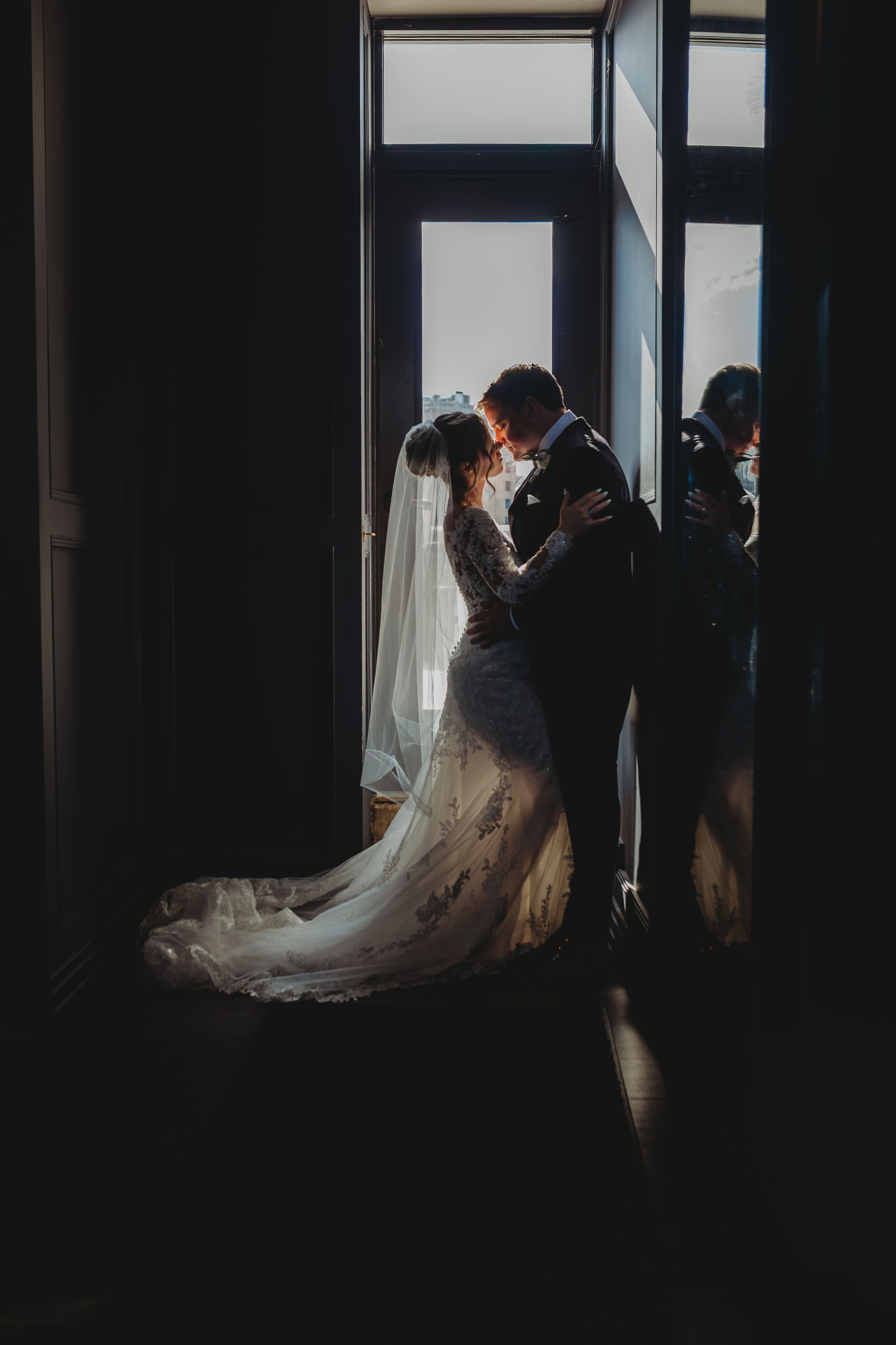 A romantic silhouette of newlyweds embracing by a window, with the bride's lace gown and long veil flowing behind her.