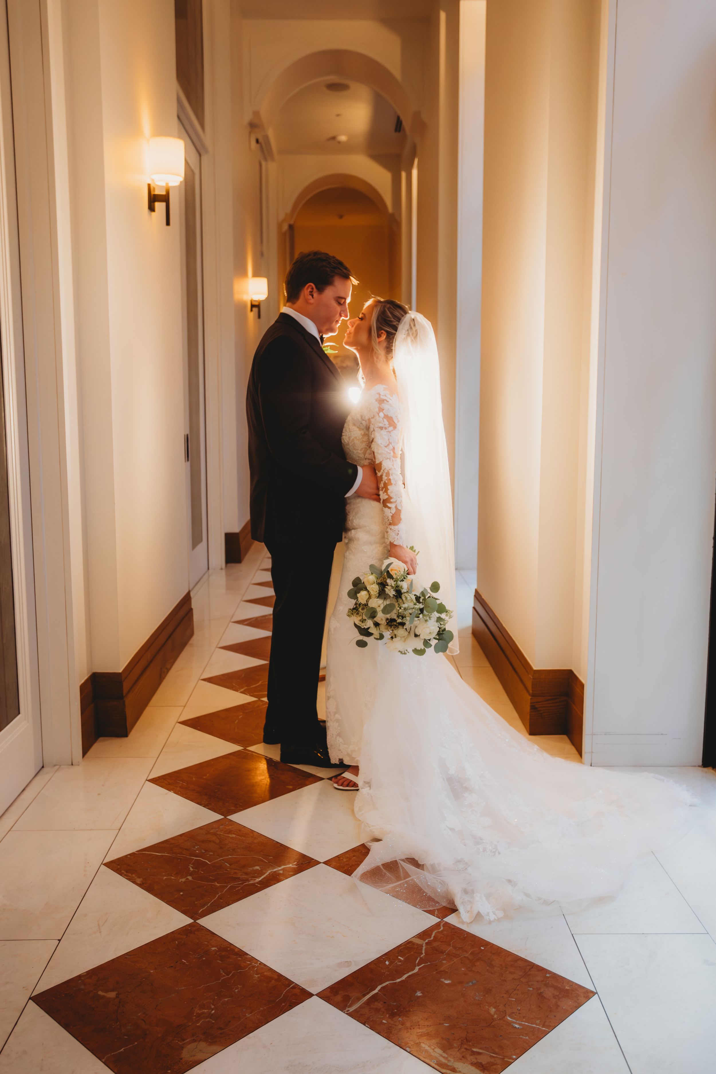 A romantic wedding photo of a couple embracing in an elegant hallway with marble checkered floors and soft lighting.