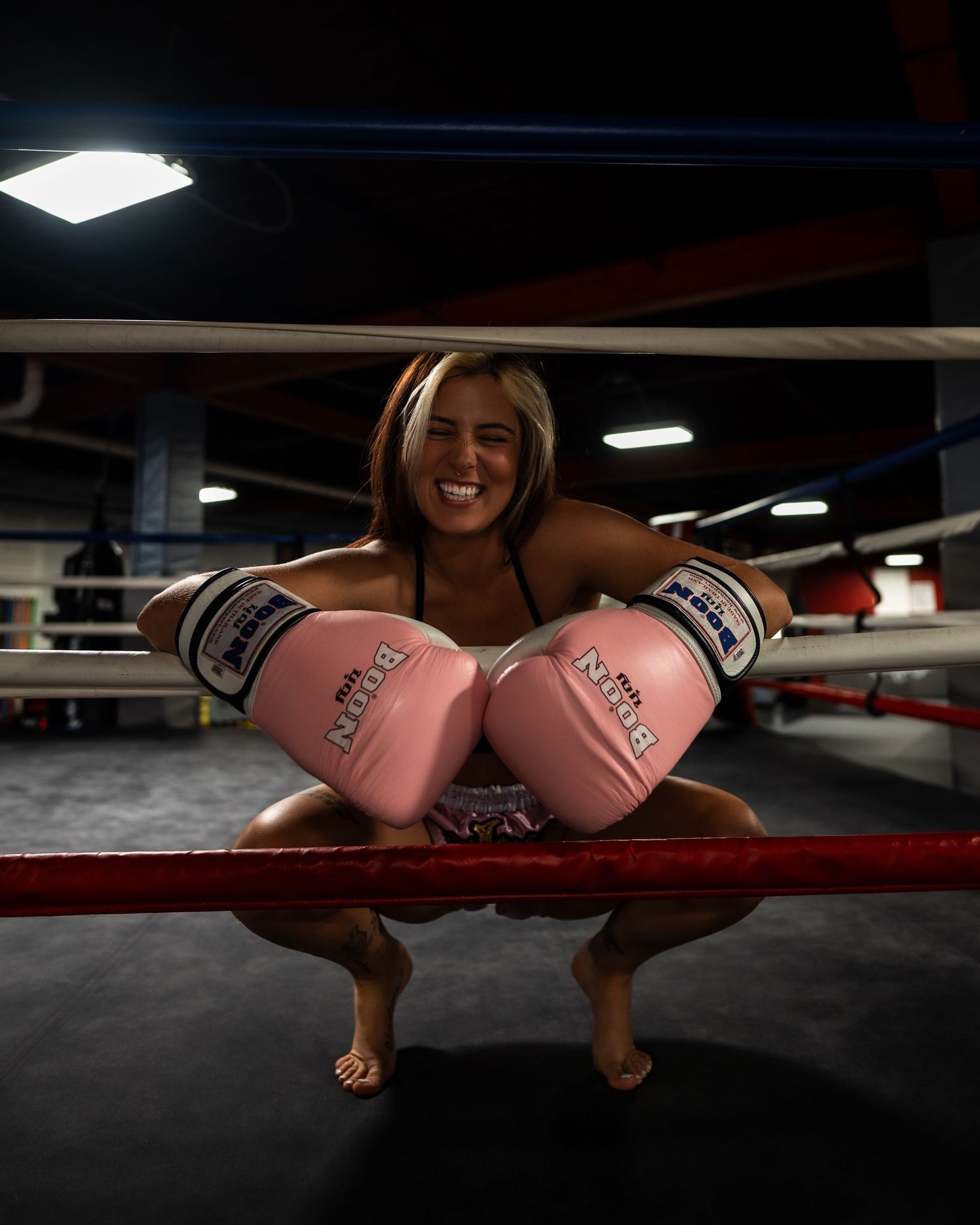 A boxer leans on boxing ring ropes wearing pink gloves while smiling in a dimly lit gym setting.