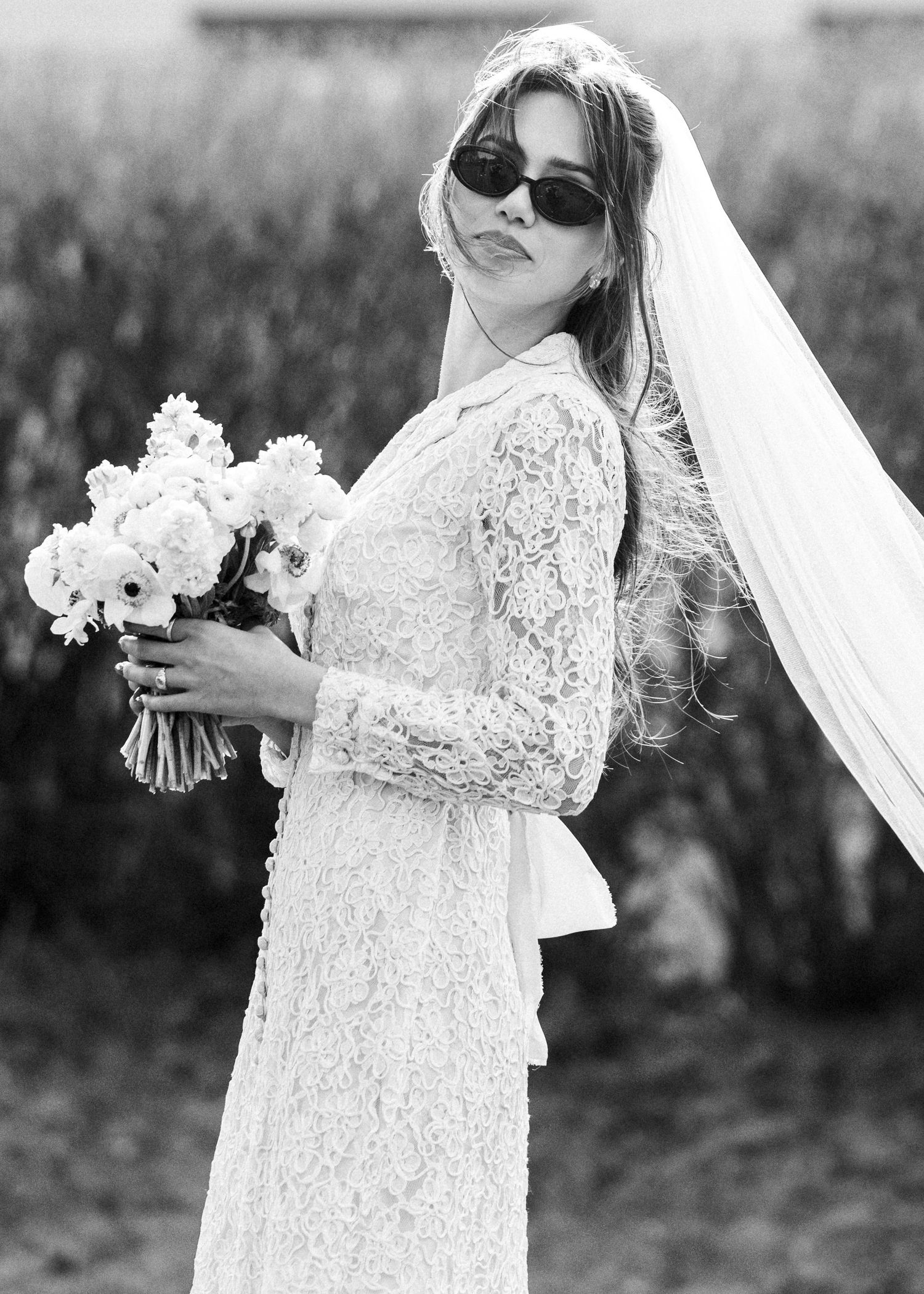 Black and white photo of a bride in a lace wedding dress, sunglasses and veil holding a white floral bouquet.