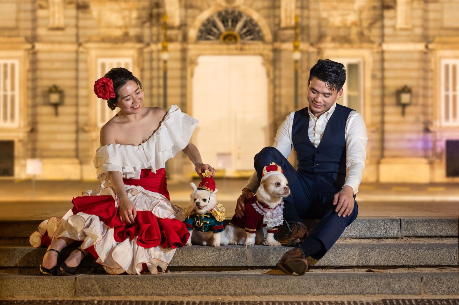 Two people in vintage-style costumes sit on stone steps with plushies in front of an elegant historic building at night.
