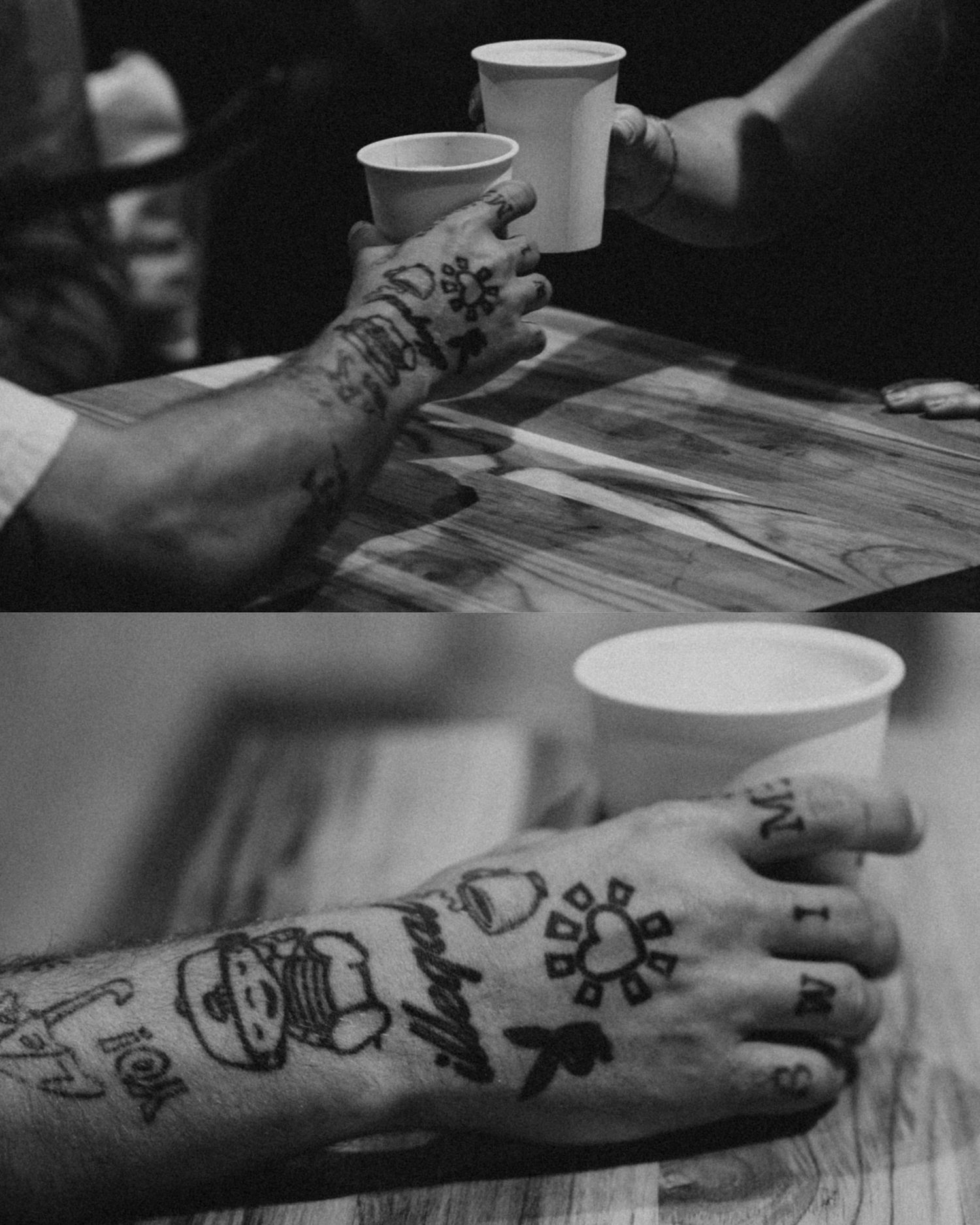 Black and white image of tattooed hands holding white disposable cups across a wooden table surface.