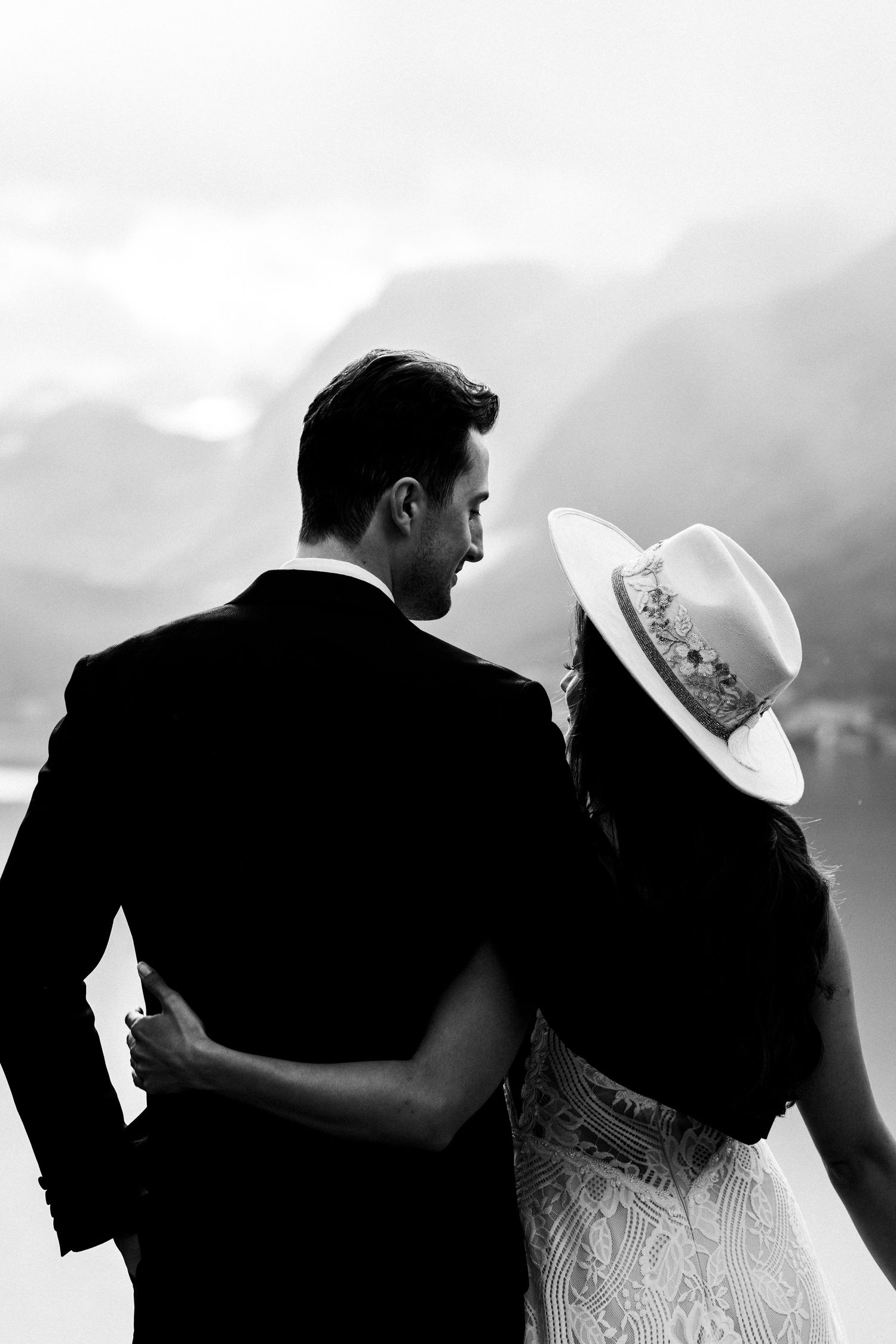Black and white photo of newlyweds embracing while looking at mountain view, bride wearing white hat and lace dress.