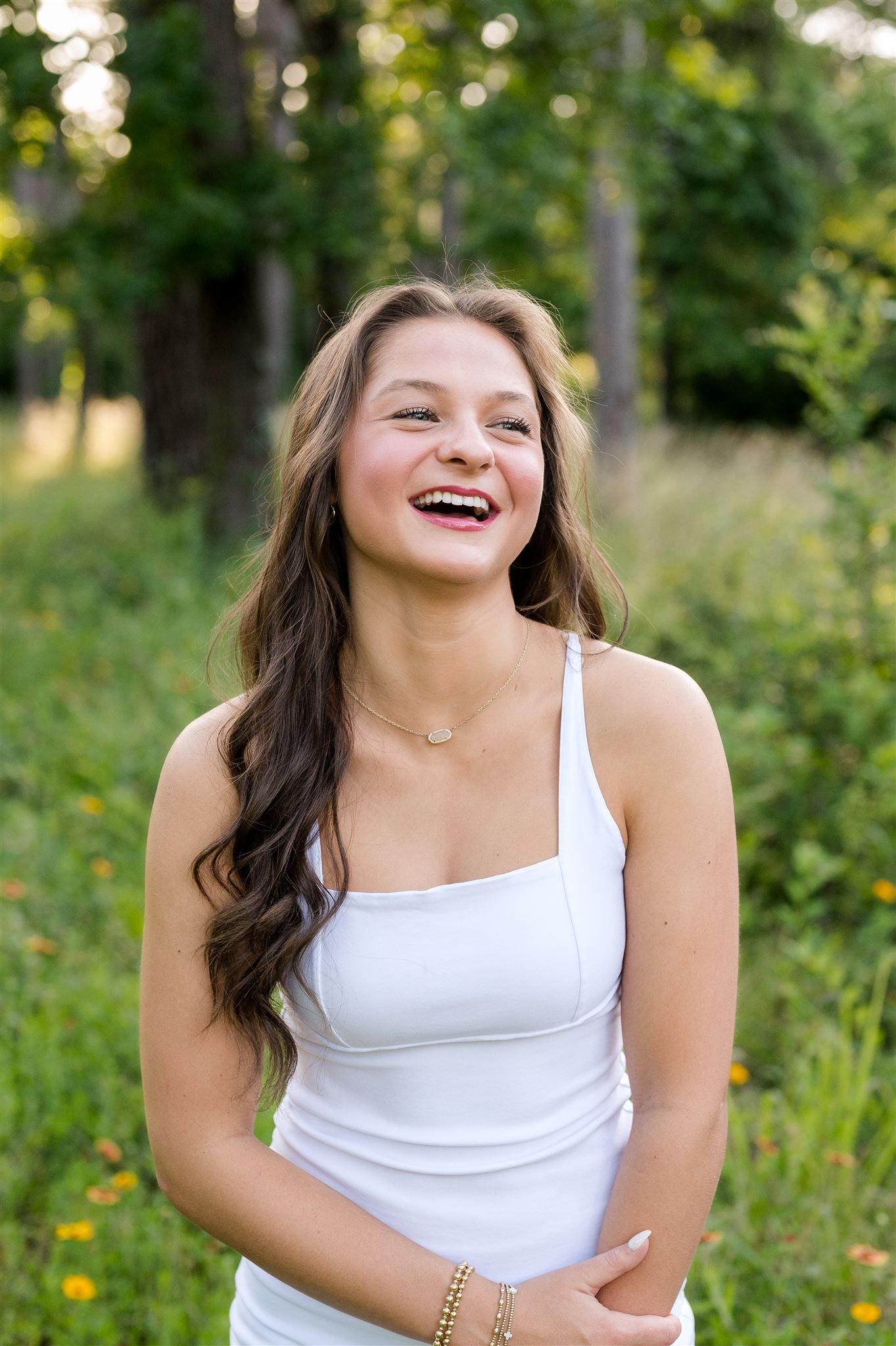 Summer portrait of someone in a white tank top laughing outdoors with sunlit greenery and trees in the background.