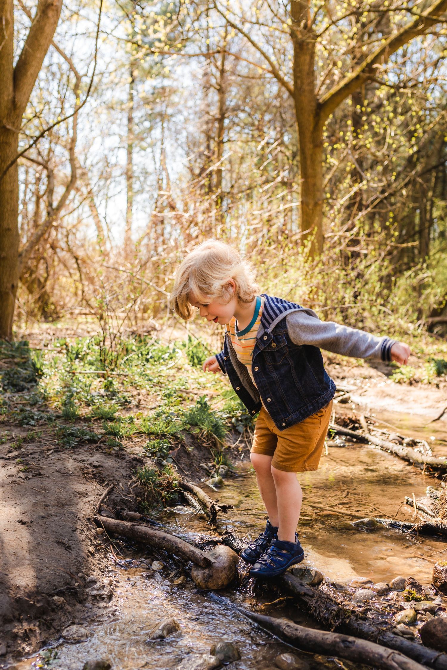 Child in blue denim vest and yellow shorts balancing on logs during sunny forest adventure on woodland trail.