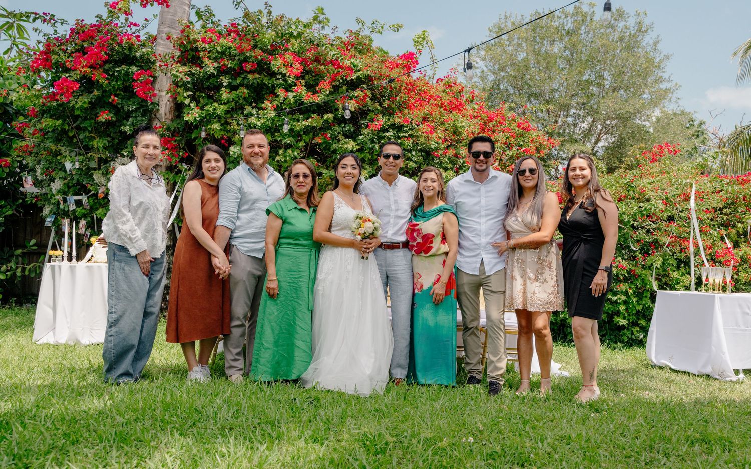 A group in formal attire poses together in front of vibrant flowering bougainvillea bushes at an outdoor garden event.