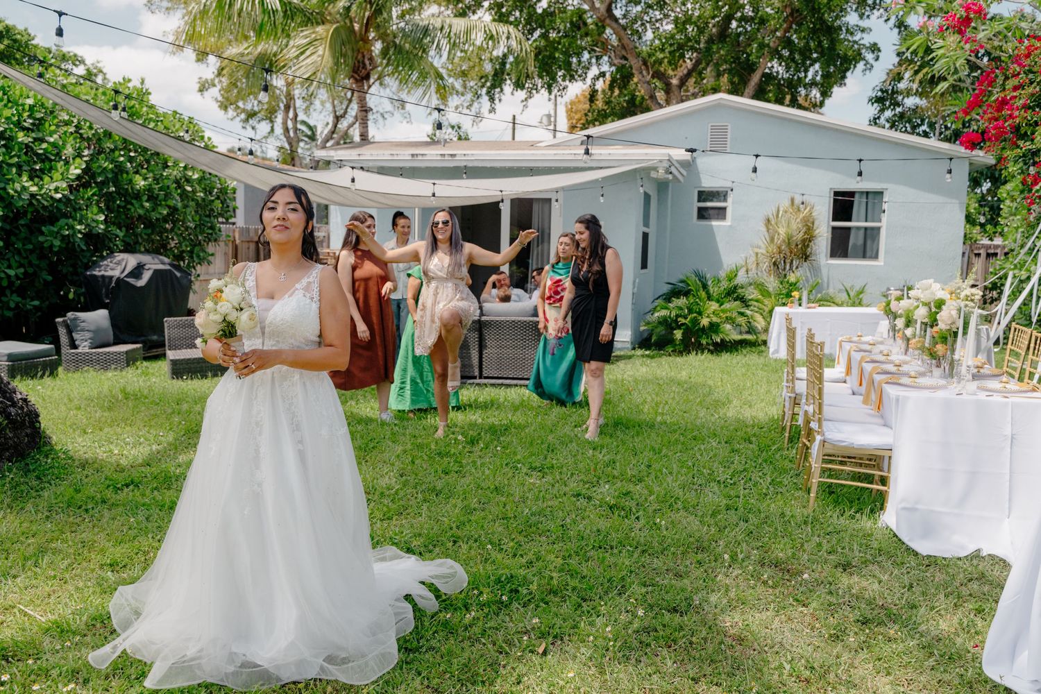 A backyard party with guests on a green lawn in front of a white house with palm trees and decor.