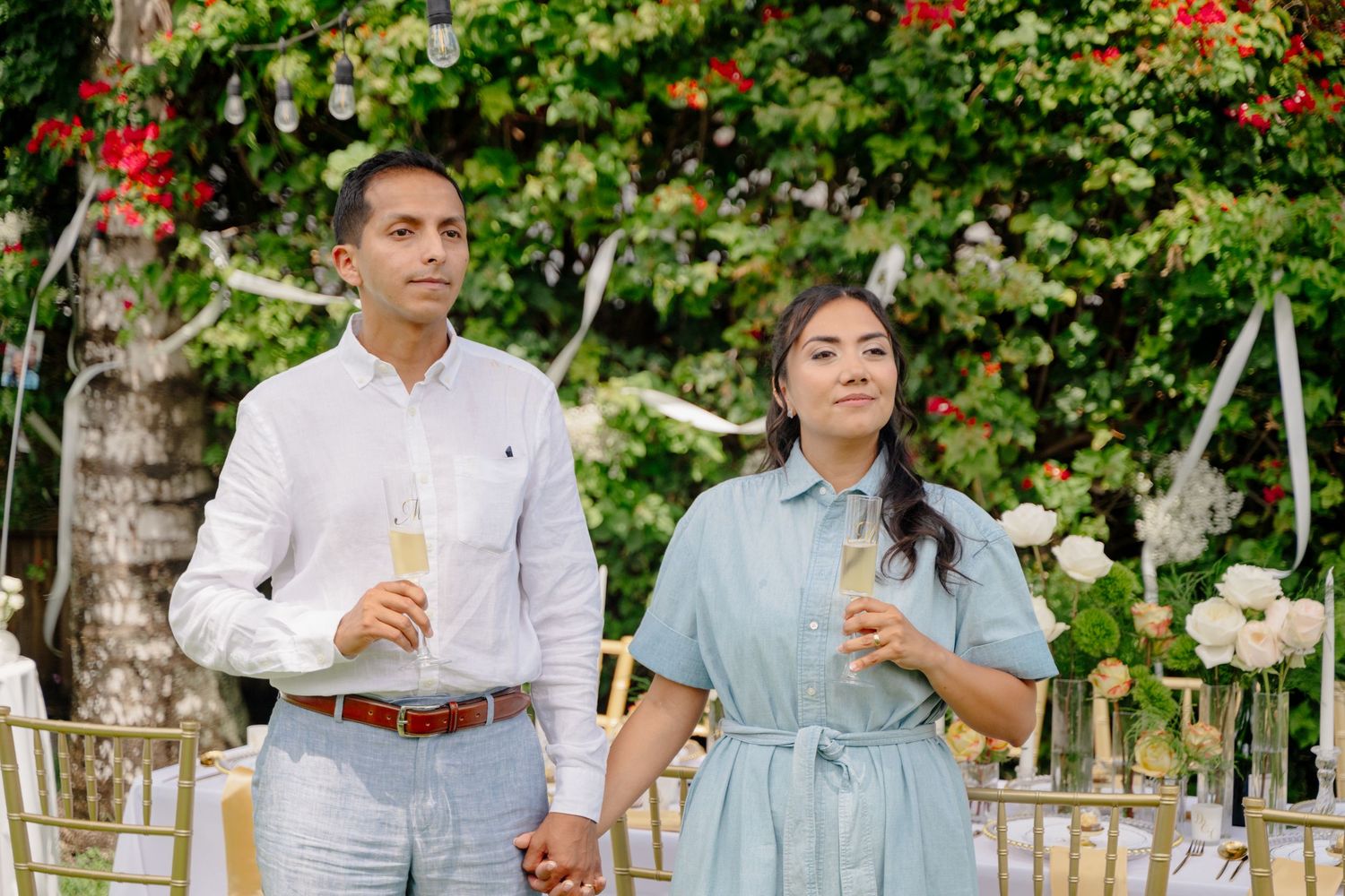 A couple in elegant attire holding beverages stands together in a garden setting with flowering bushes and decorative chairs.