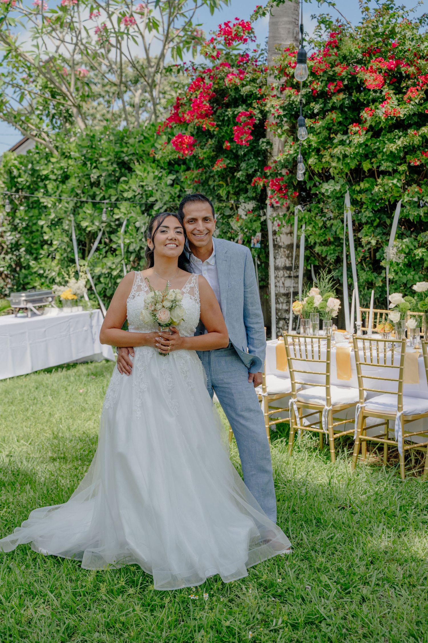A wedding couple poses in a garden setting with red flowers and gold chairs arranged for an outdoor ceremony.