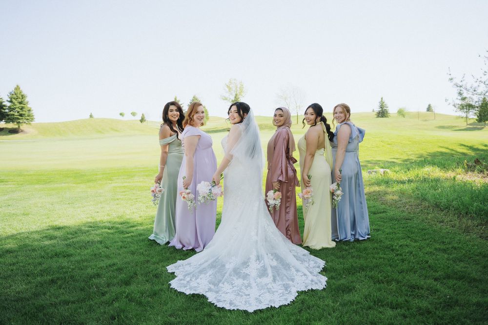 Wedding party in pastel bridesmaid dresses lounging on sunny grassy hillside during outdoor photoshoot.
