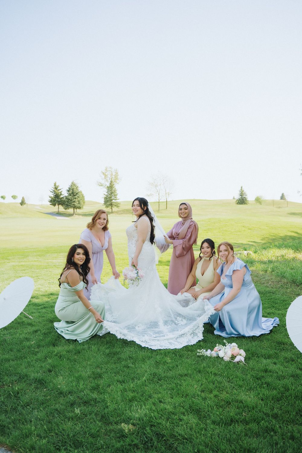 Bridesmaids in soft colored dresses sitting together on green lawn with bridal gown spread out.