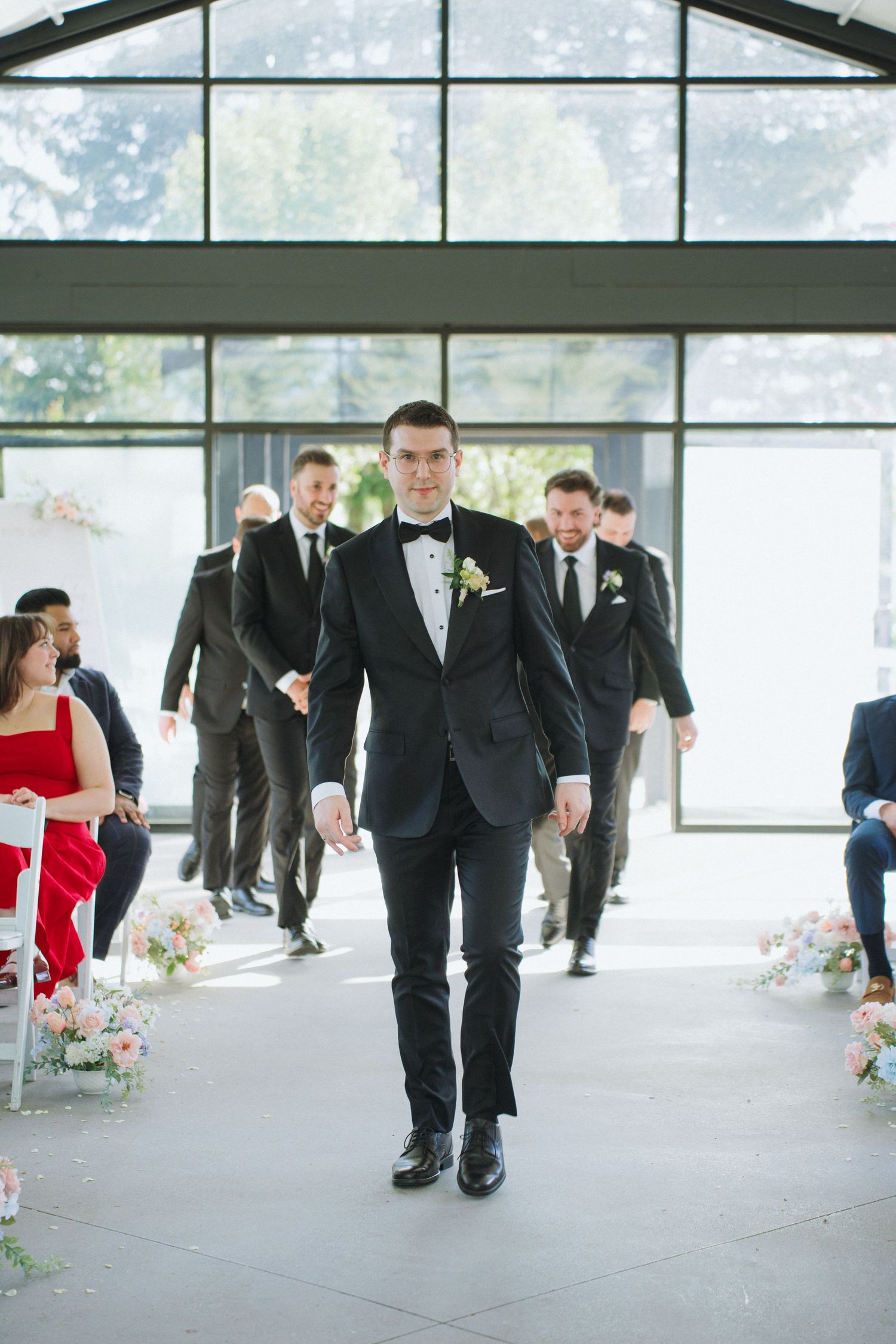 Wedding party members in formal attire walking through modern venue with floor-to-ceiling windows.