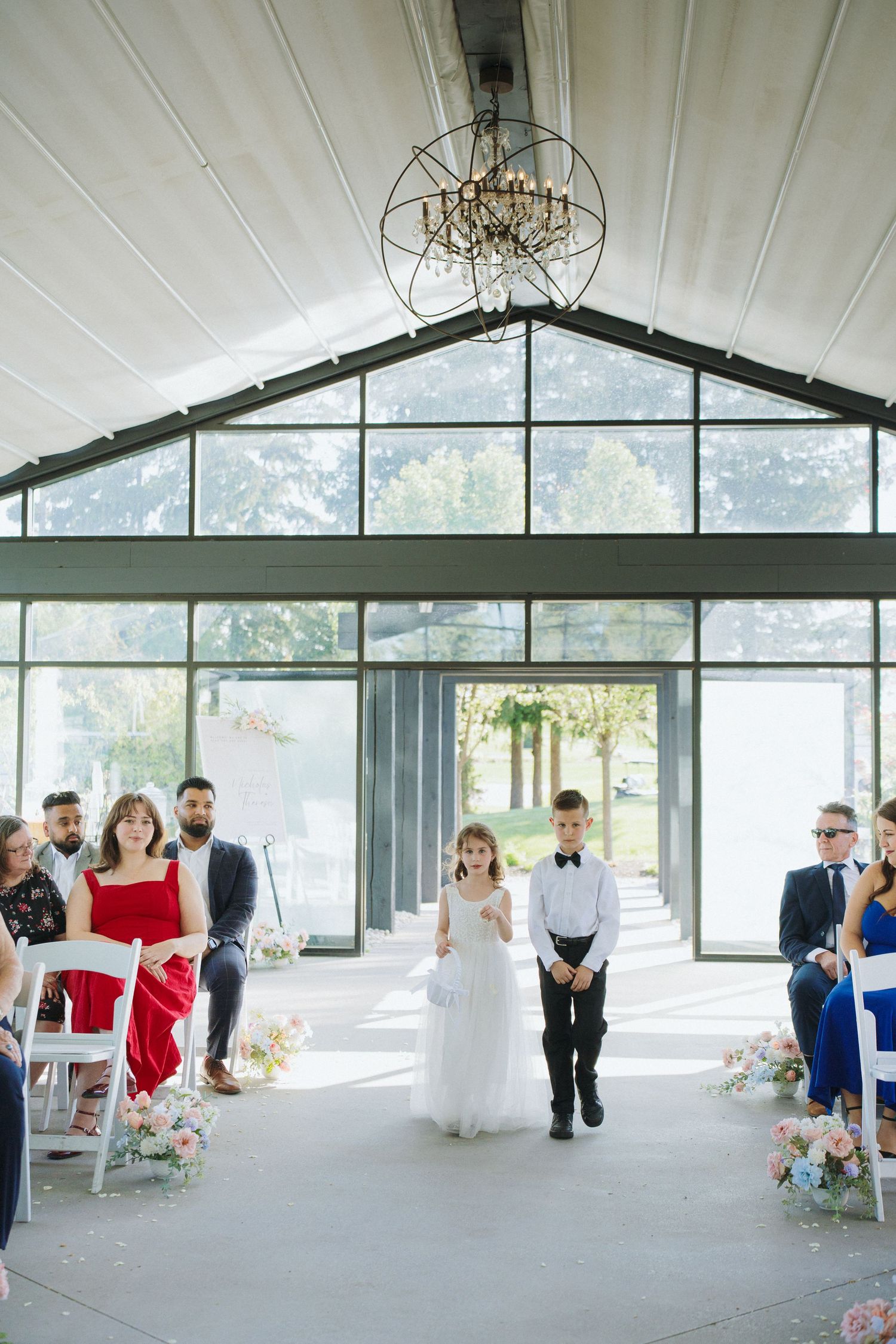 Modern wedding venue interior with white ceiling, chandeliers and large windows overlooking trees.