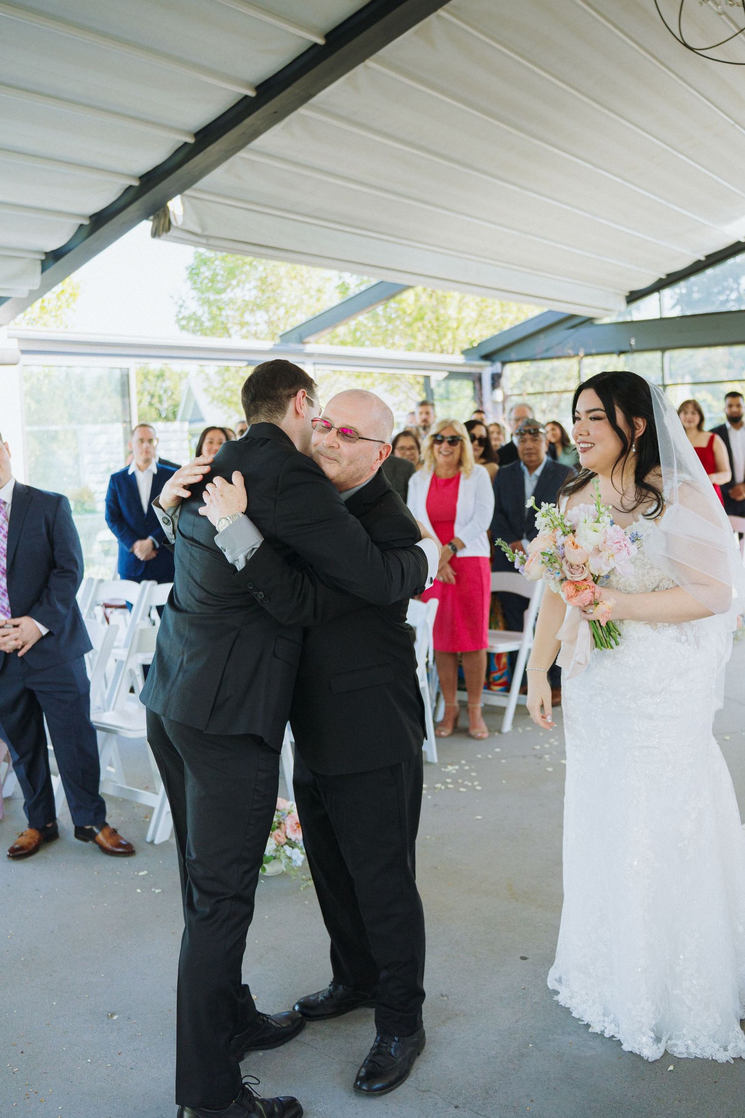 Wedding ceremony moment with two men embracing as bride looks on in an elegant covered outdoor venue.