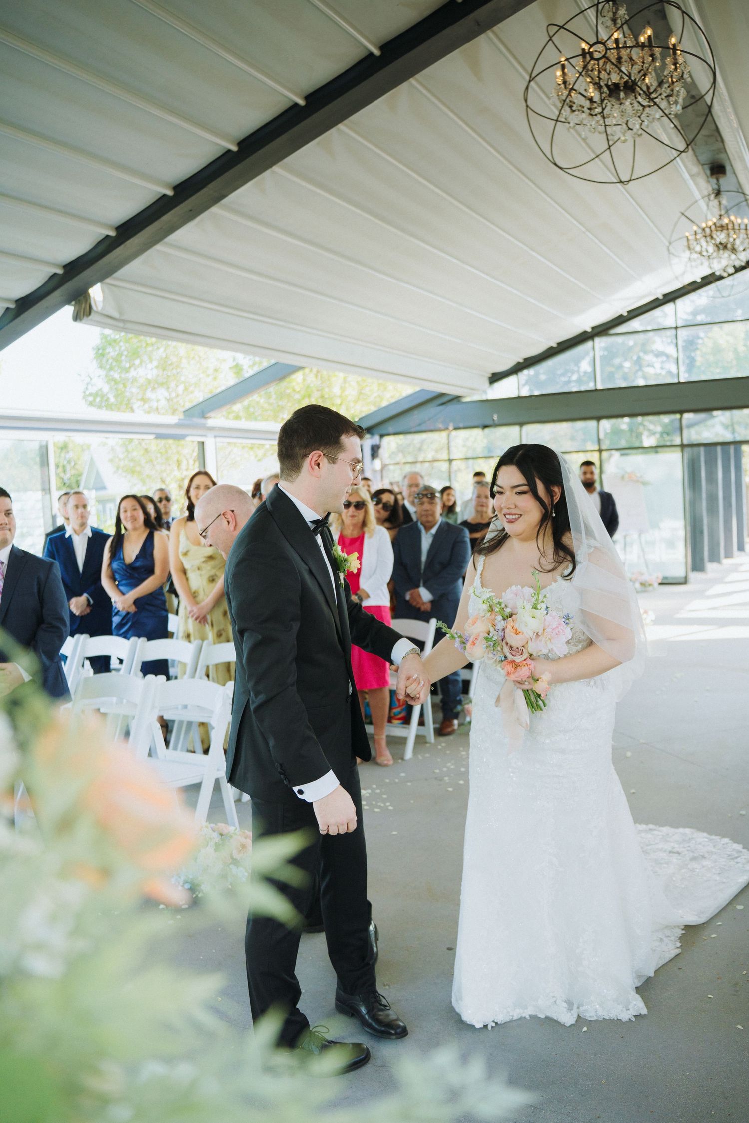 Couple exchanges vows during wedding ceremony in bright indoor-outdoor space with chandelier.