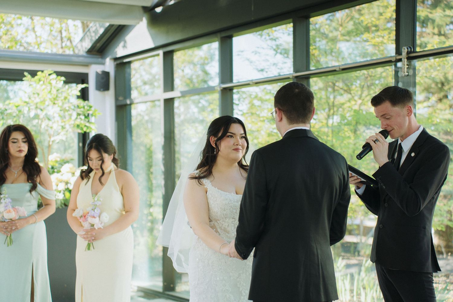 Wedding ceremony underway in modern glass-walled venue with bridal party standing at altar.