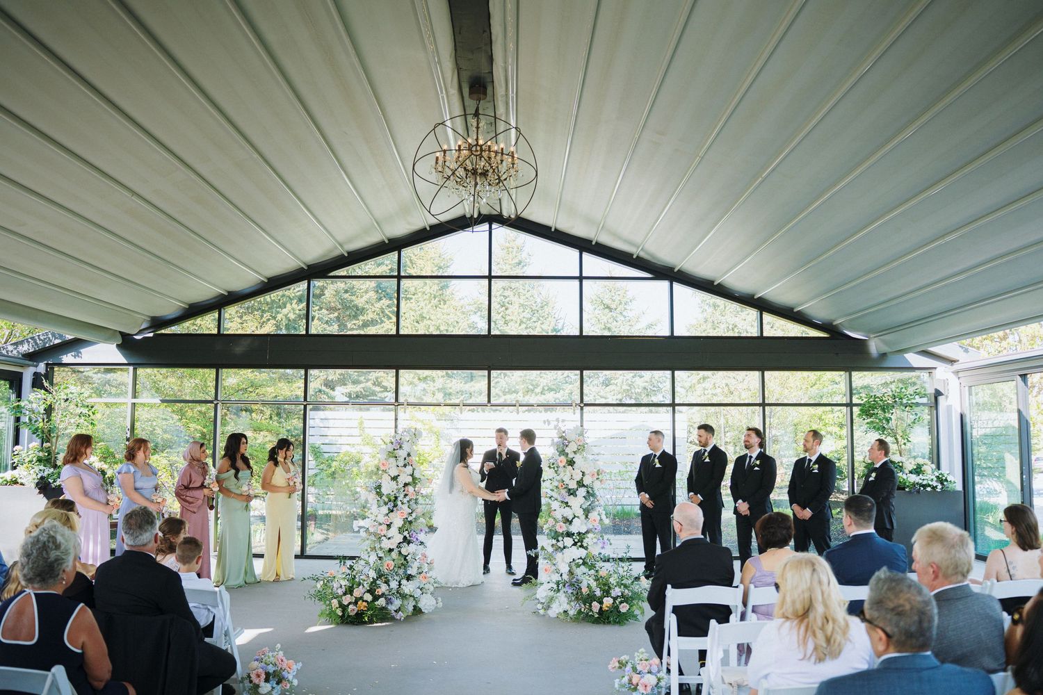 Wide view of wedding ceremony in modern pavilion with white ceiling and glass walls.