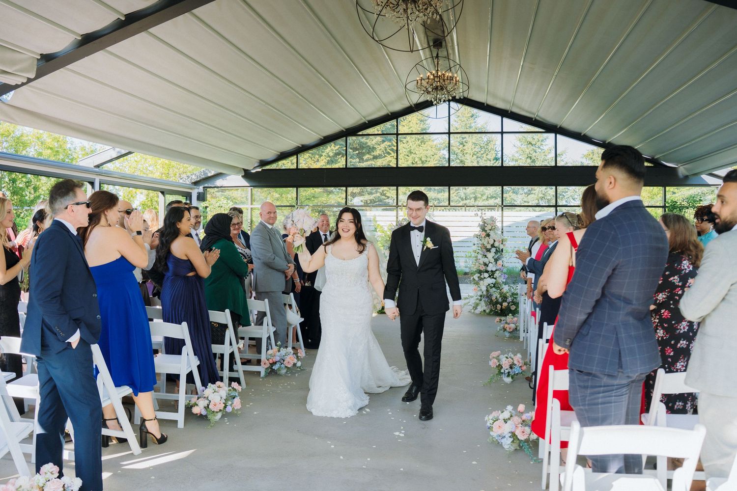 Newlyweds walk down aisle after ceremony in glass pavilion decorated with white flowers.