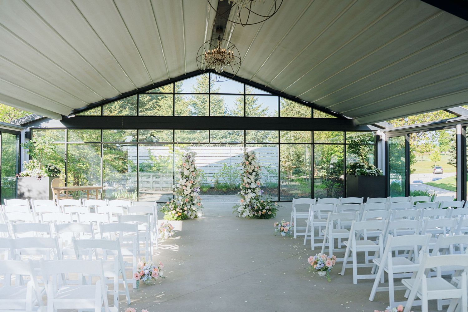Empty wedding venue with white chairs and floral arrangements in modern glass-walled pavilion.