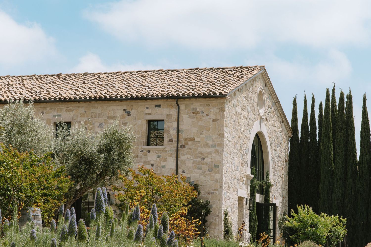 Mediterranean stone villa with arched entryway surrounded by cypress trees and flowering gardens against blue sky.
