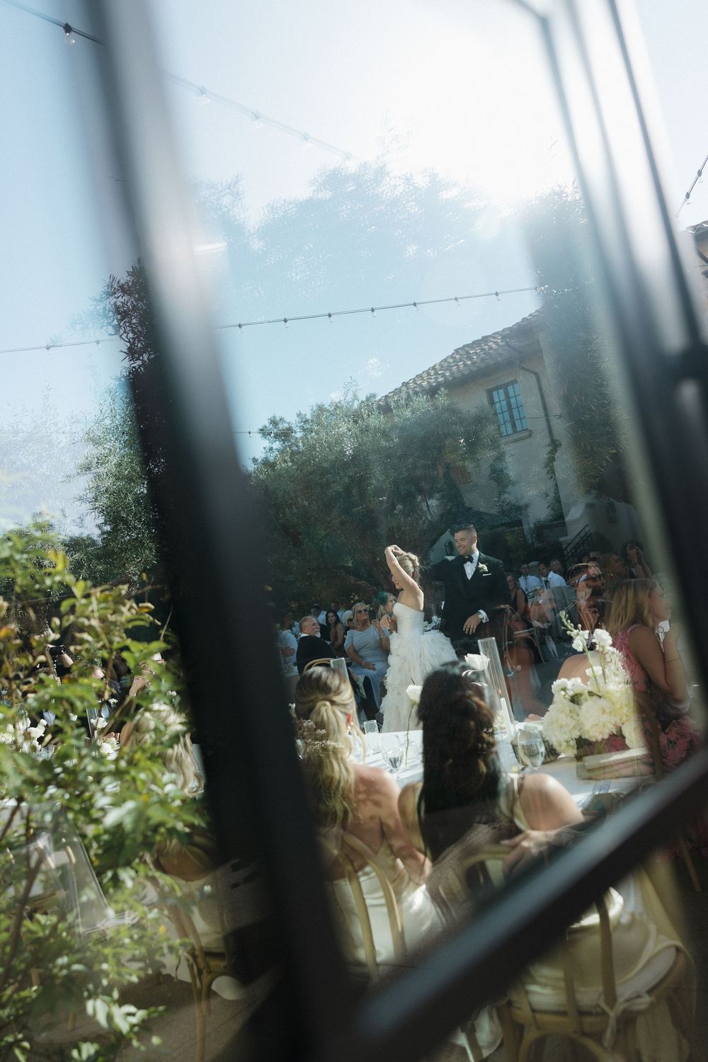 Artistic wedding ceremony view through window panes showing couple and guests gathered at an outdoor venue.