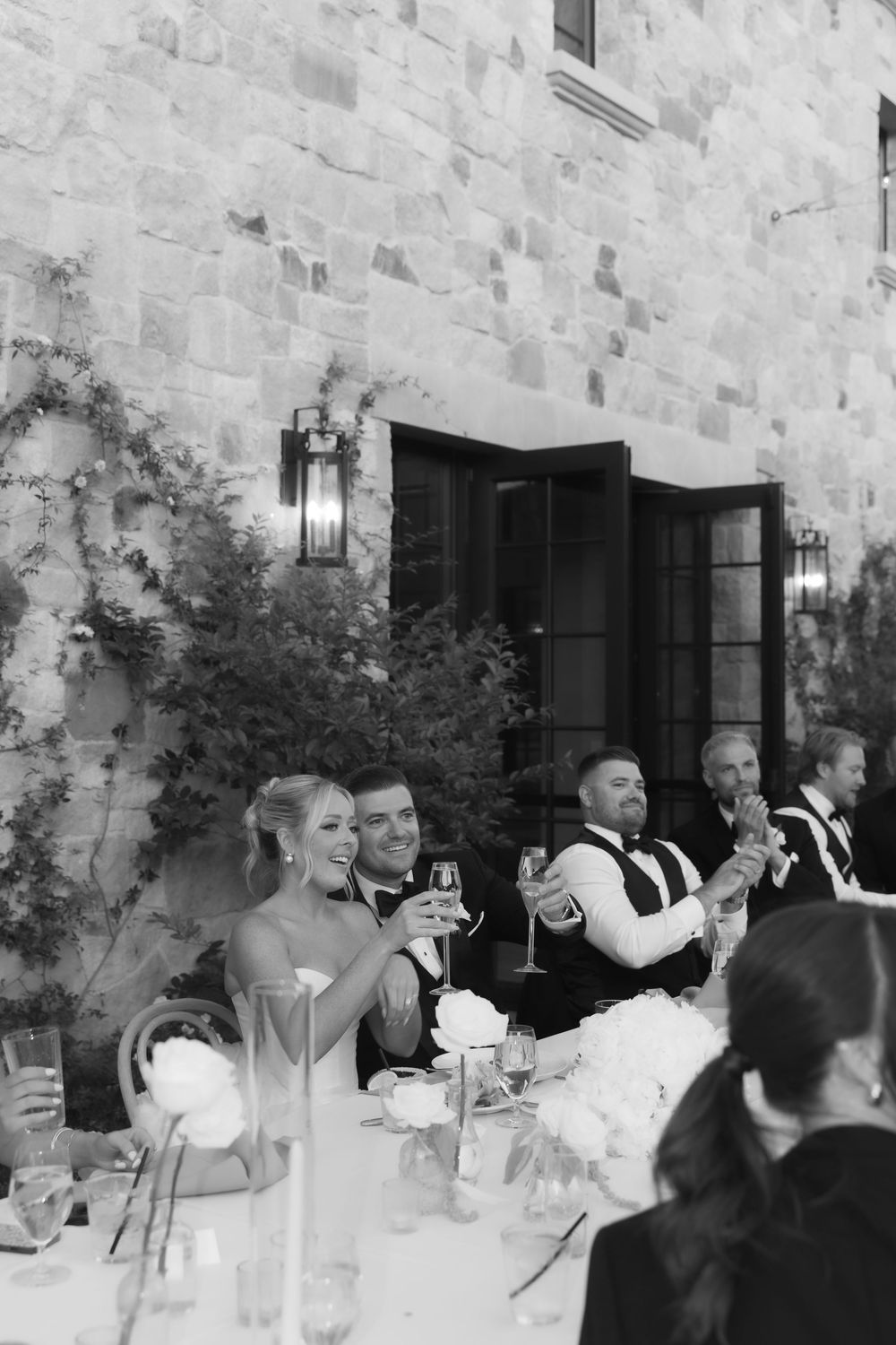 Wedding guests enjoying dinner and drinks at a rustic stone-walled venue with elegant black and white formal attire.