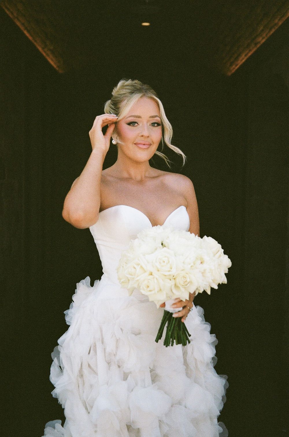 Bride in strapless white ruffled wedding gown holds elegant bouquet of white roses against dark background.