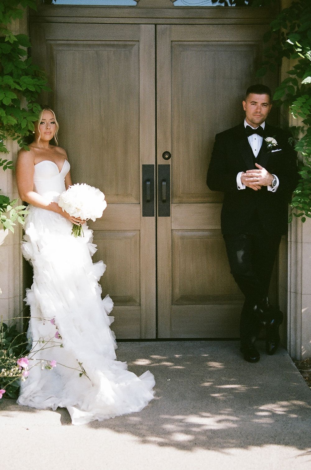 A bride in a white strapless gown and groom in black tuxedo stand on opposite sides of elegant wooden double doors.