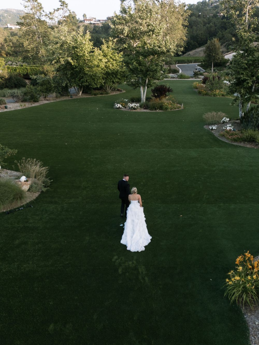 An aerial view of a wedding couple standing on a manicured lawn surrounded by landscaped gardens.