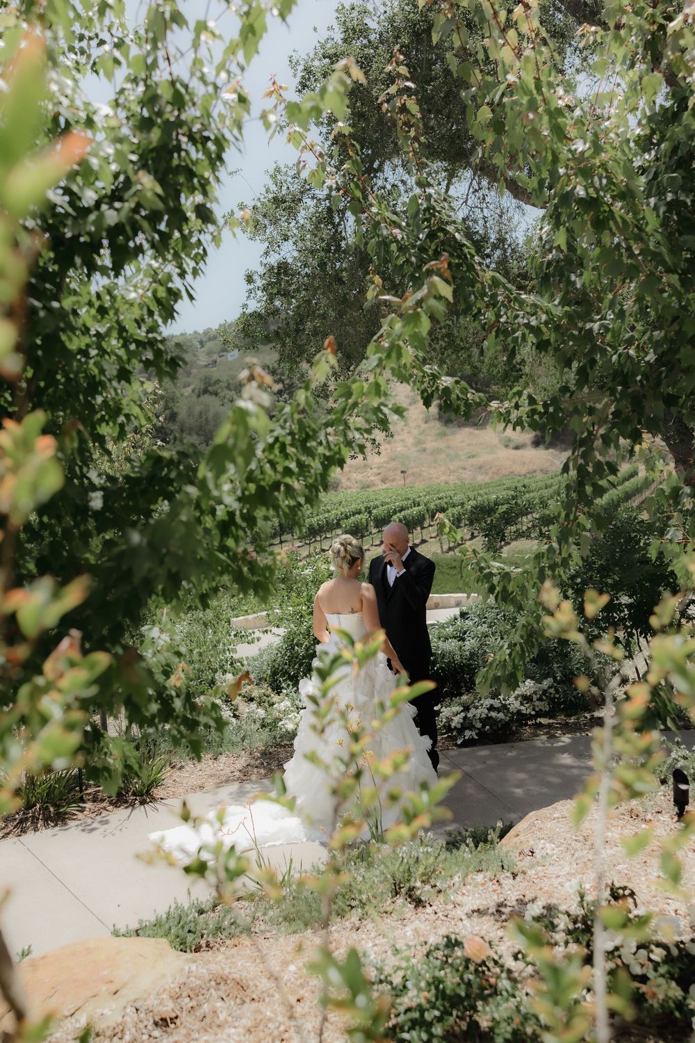 Wedding couple walking along garden path surrounded by lush green trees and foliage.