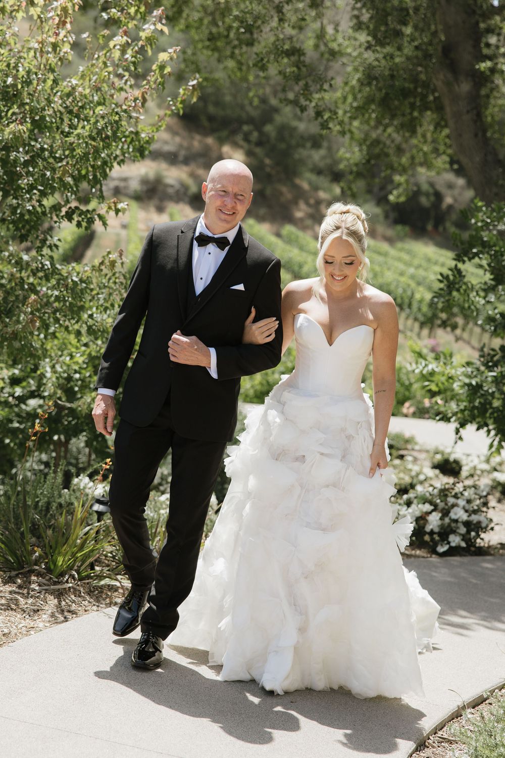 Newlyweds walking together on garden path, bride in flowing white gown and groom in classic tuxedo.