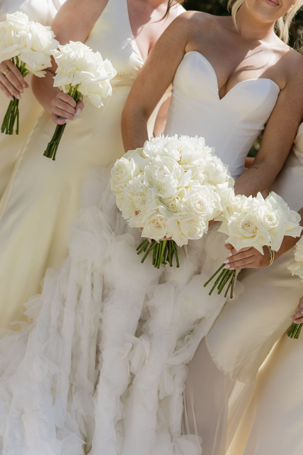 Elegant white wedding bouquets held against flowing white wedding dresses in bright sunlight.
