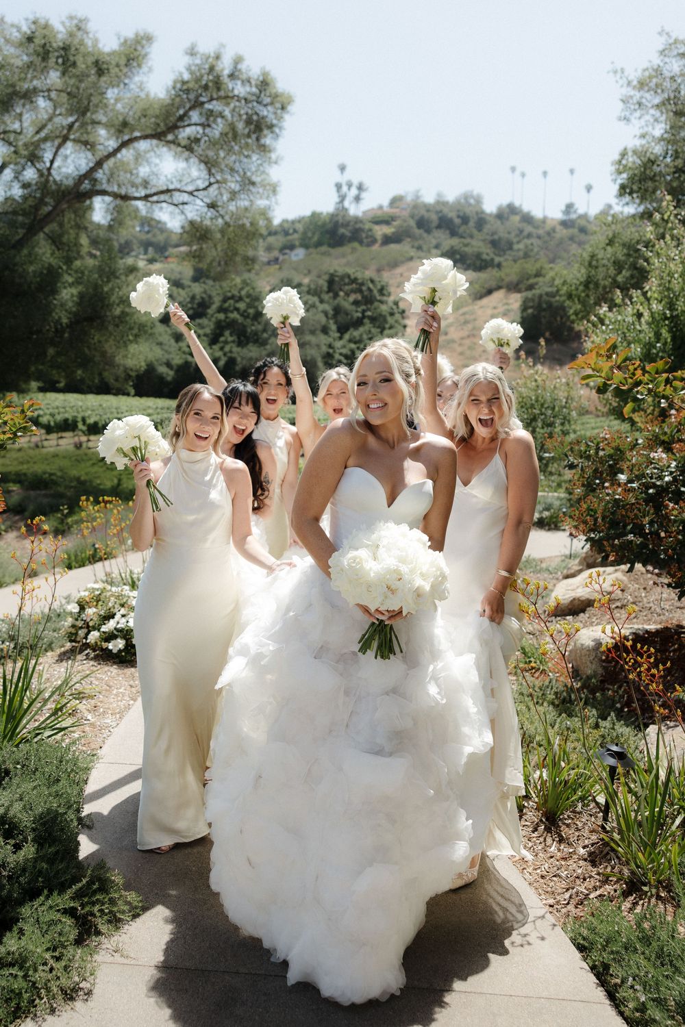 Bridal party dressed in white gowns walk along garden path holding white floral bouquets on a sunny day.