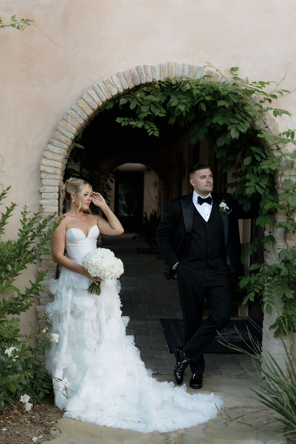 A couple in wedding attire pose in front of an arched stone doorway with climbing vines at an elegant venue.