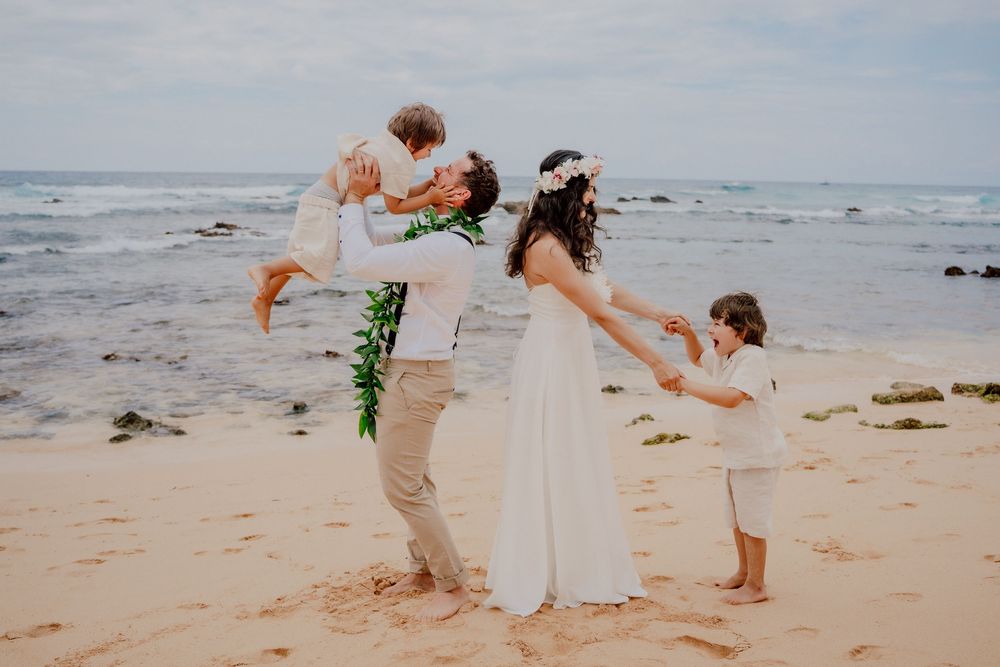 A joyful family plays and lifts their children up while celebrating on a sandy beach in Hawaii.