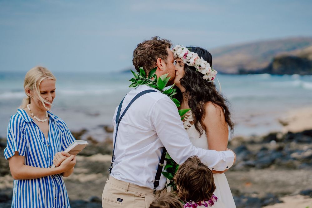 A beachside wedding ceremony takes place on a rocky shore with mountains in the background and blue ocean waves.