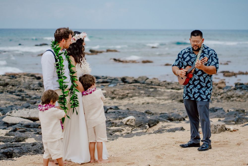 A musician plays ukulele on a rocky beach while others in traditional Hawaiian leis stand on the shore.