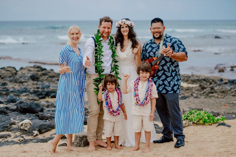 Family group wearing festive Hawaiian leis poses together on rocky beach with ocean in background.
