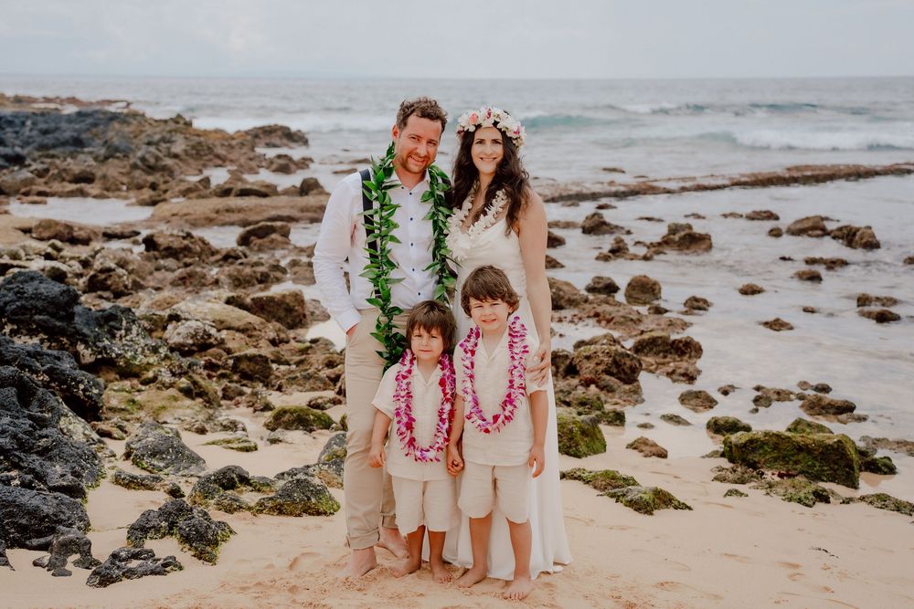 A family wearing traditional Hawaiian leis poses together on a rocky beach shoreline at sunset.