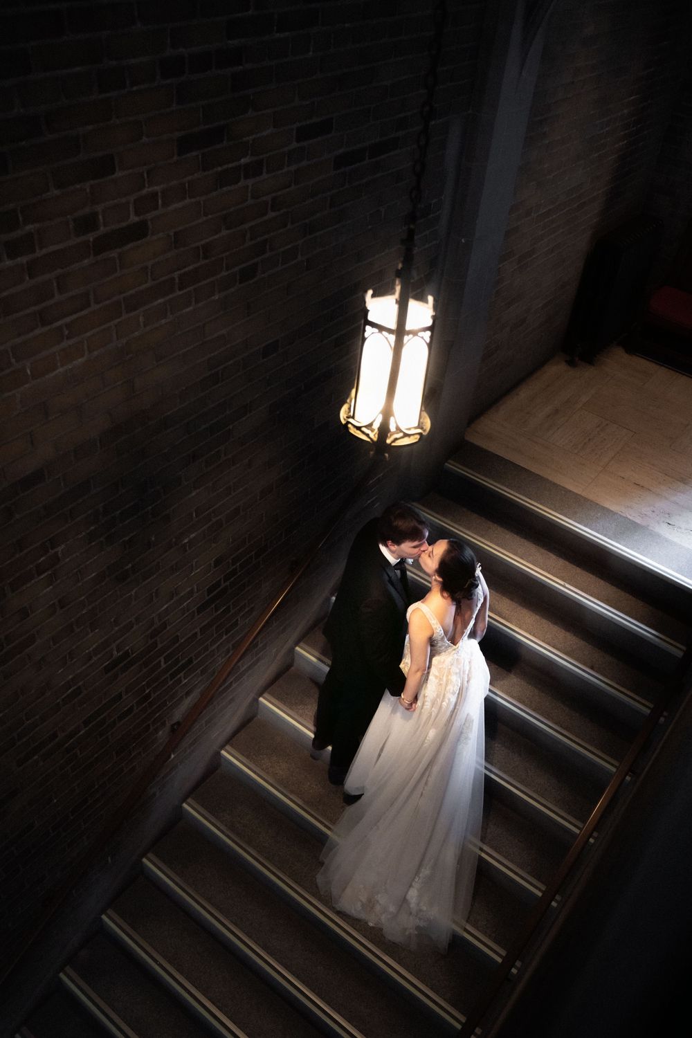 Newlyweds share intimate moment on dimly lit staircase with vintage pendant light.