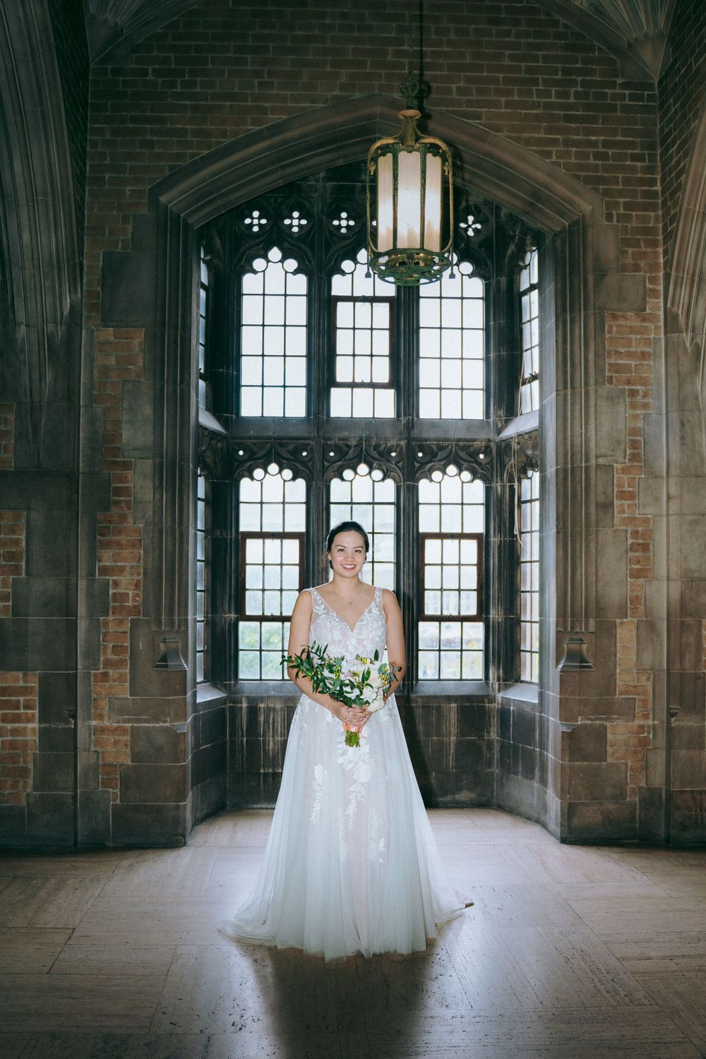 Bride holds bouquet in gothic hall with stunning arched windows and architectural details.